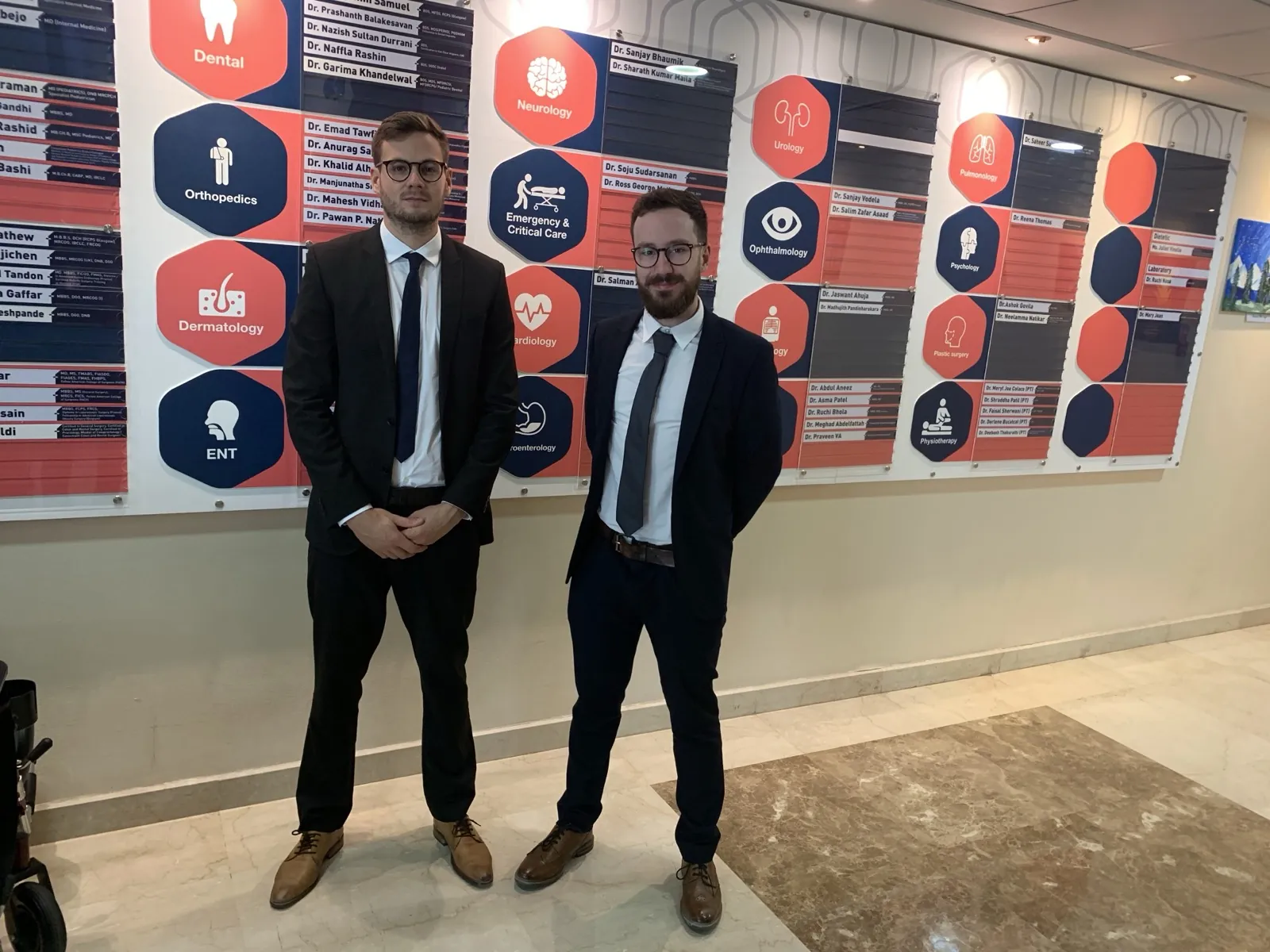 Ollie Capel and Louis Meletiou of Medico Digital standing in a hospital lobby in front of a wall of department signs